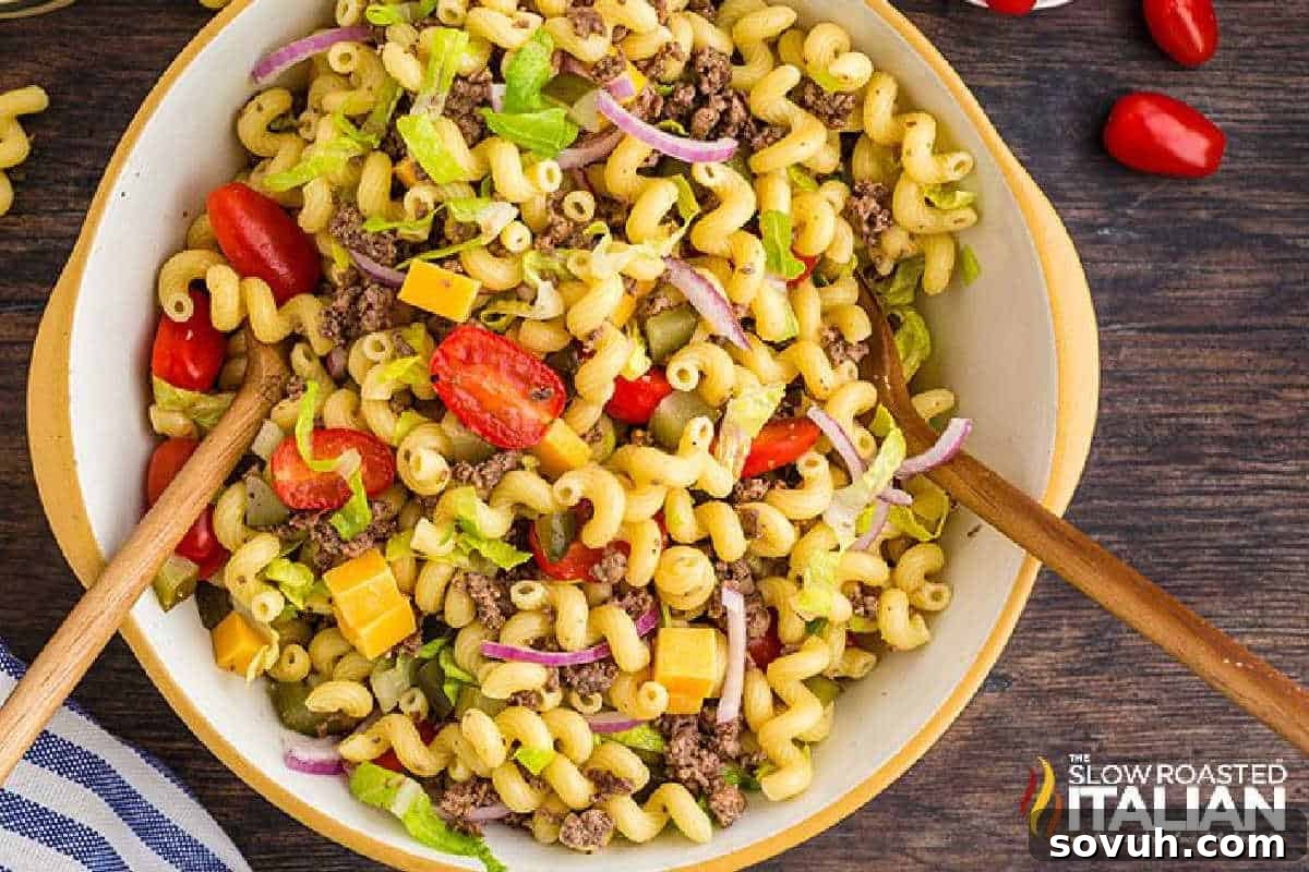 A bowl of macaroni salad with ground beef, cherry tomatoes, red onions, and diced cheddar cheese, served with wooden spoons.