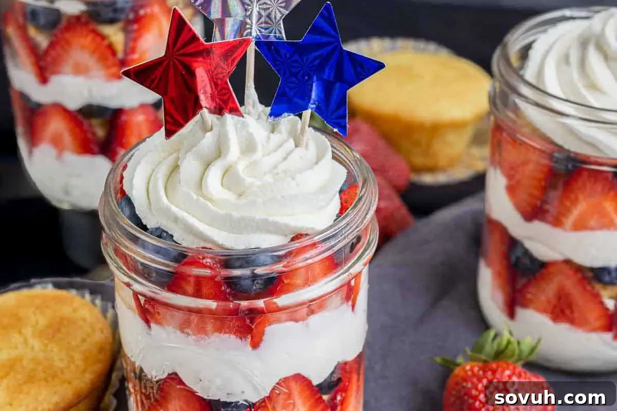 Layered dessert in mason jars with whipped cream, strawberries, blueberries, and decorative red and blue star toppers. A plain cupcake and fresh strawberries are visible in the background.
