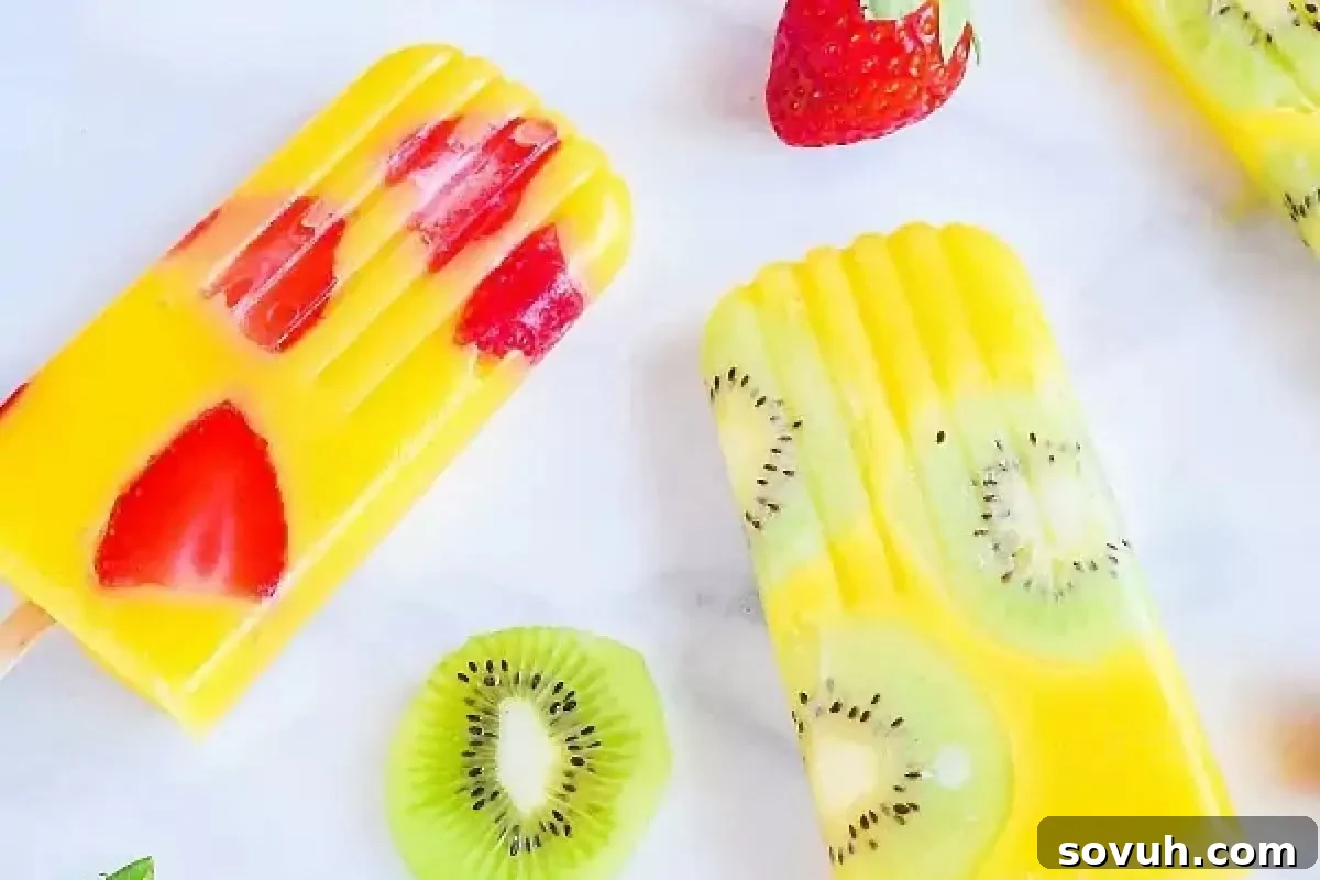 Two popsicles, one with strawberry pieces and one with kiwi slices, are lying on a white surface. There are additional whole and sliced strawberries and kiwis around them.