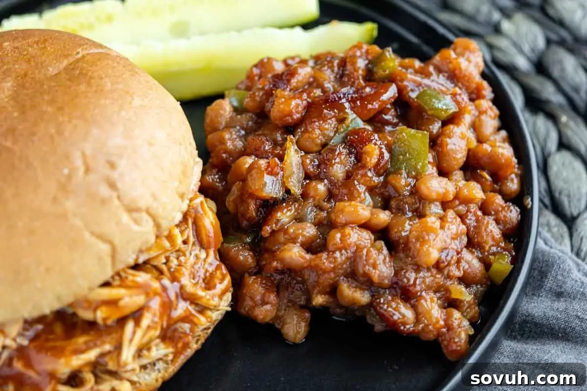 Close-up of a plate with a pulled chicken sandwich on a bun, a side of Baked Beans Recipe with diced green peppers, and pickle spears in the background.