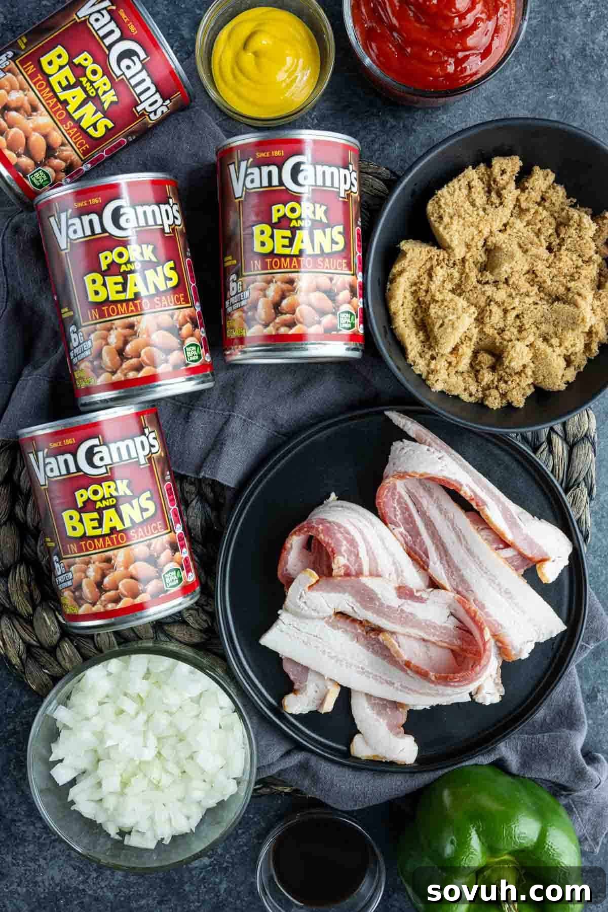 An arrangement of ingredients includes Van Camp's Pork and Beans, bacon slices, brown sugar, chopped onions, mustard, ketchup, and a green bell pepper on a dark countertop to make Baked Beans Recipe