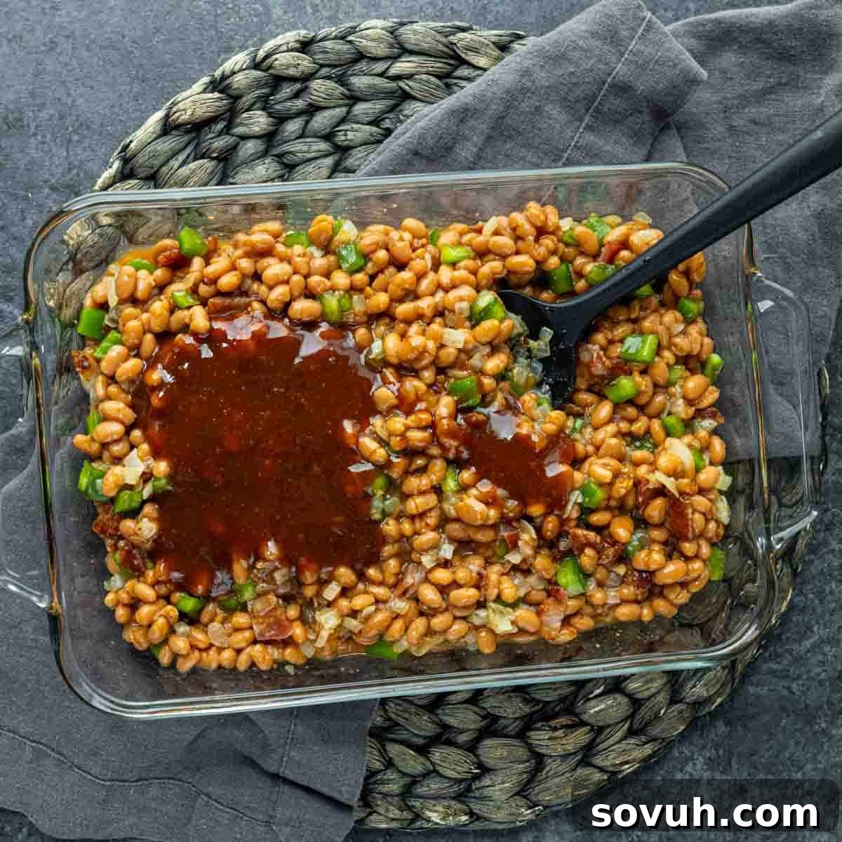 A glass dish filled with Baked Beans Recipe, chopped green bell peppers, and onions, placed on a woven mat with a gray cloth underneath. A black spoon is inserted into the baked beans.