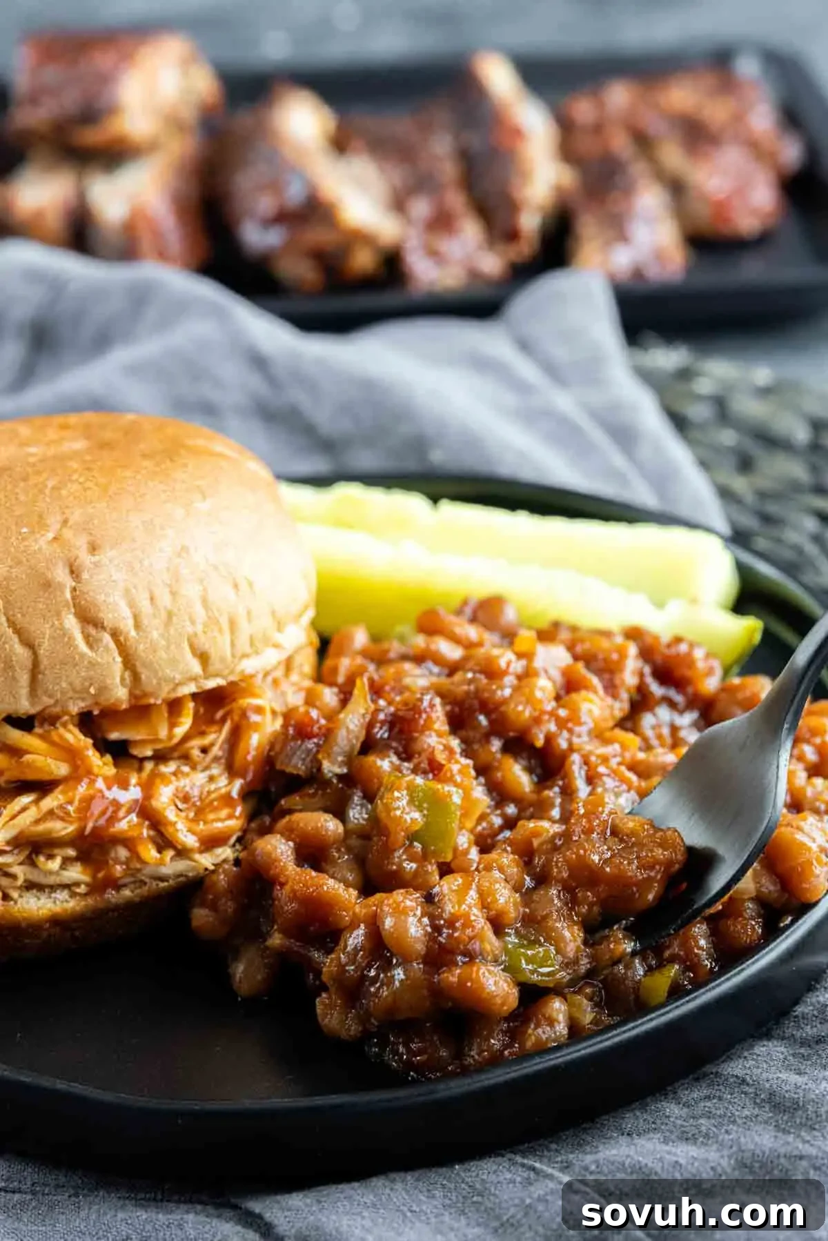 A pulled chicken sandwich with a side of Baked Beans Recipe and pickle spears on a black plate. BBQ ribs are visible in the background.