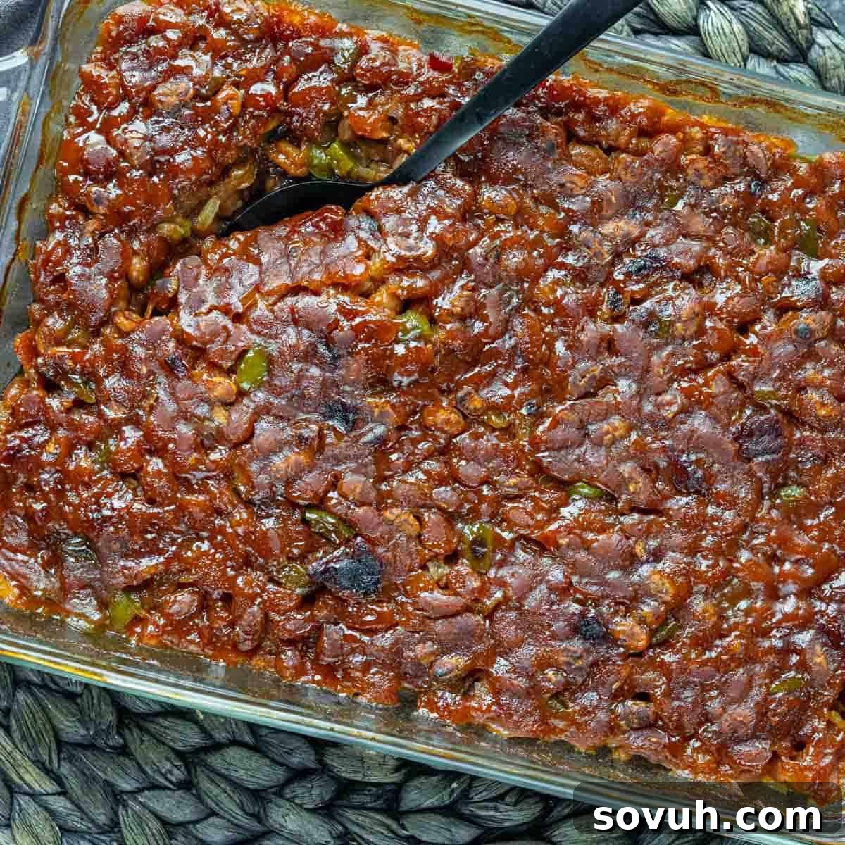 A glass baking dish filled with Baked Beans Recipe topped with crispy brown caramelized pieces, with a serving spoon inserted in one corner.