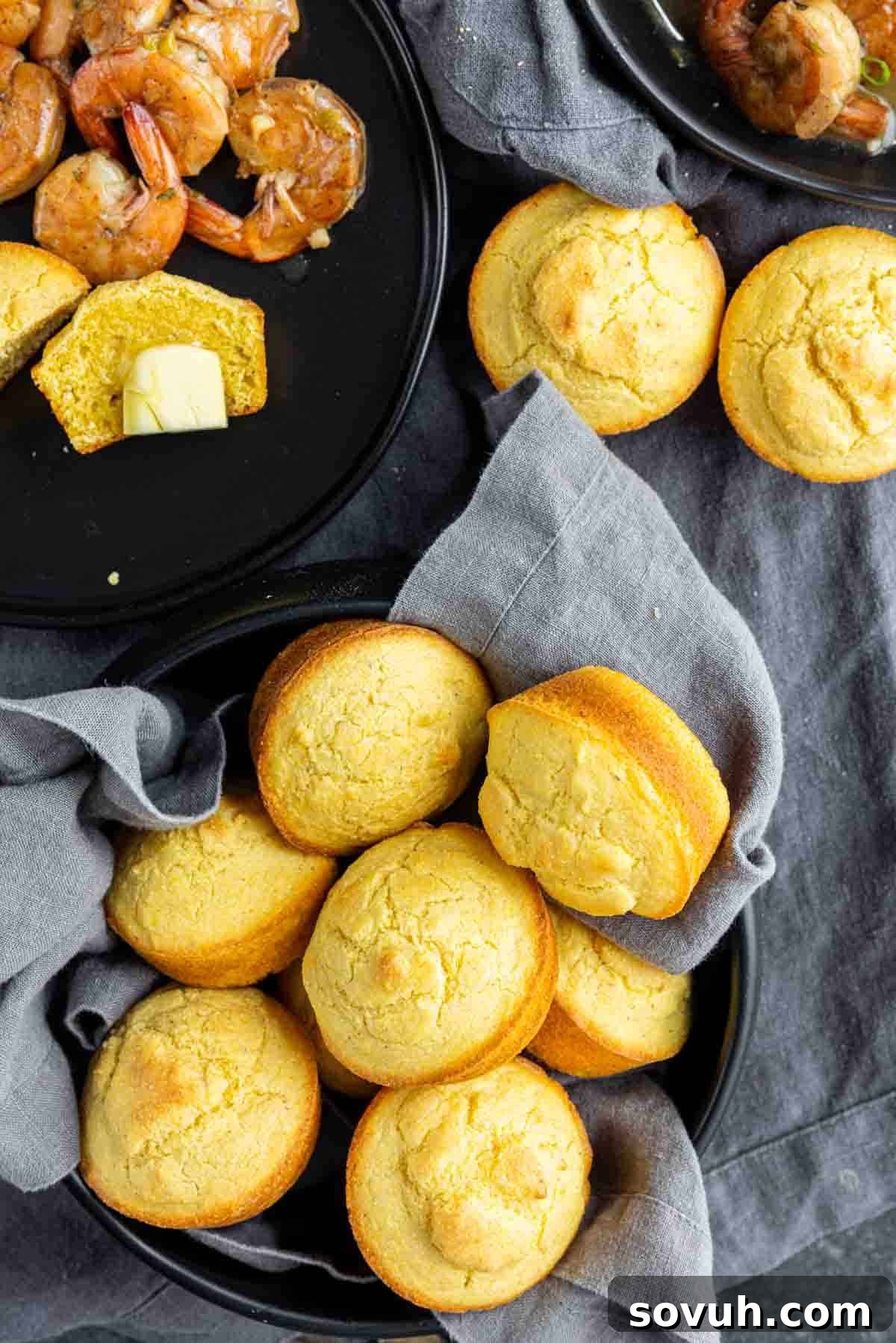 A basket of freshly baked Southern Cornbread muffins on a grey cloth, with a plate of shrimp and a buttered cornbread muffin in the background, highlighting perfect serving suggestions.