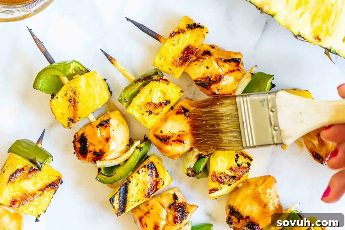 Grilling skewers with pineapple, bell peppers, and chicken are being brushed with a marinade or glaze.