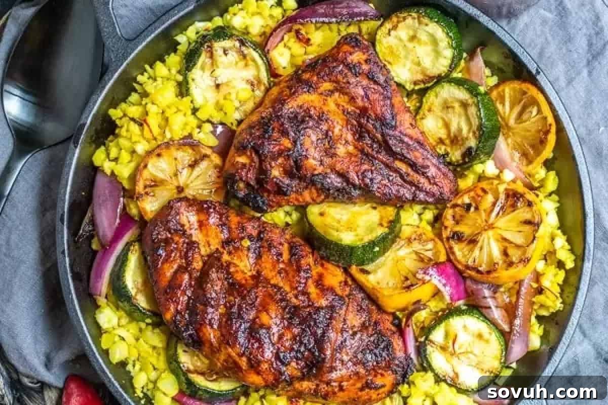 A skillet containing two pieces of grilled chicken, sliced zucchini, lemon slices, red onions, and yellow rice.