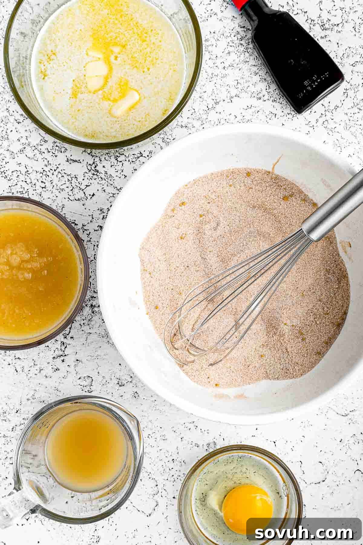 Close-up of a large mixing bowl where wet and dry ingredients are being combined to form the Apple Spice Cake batter. A whisk is visible, ensuring a smooth and consistent mixture.