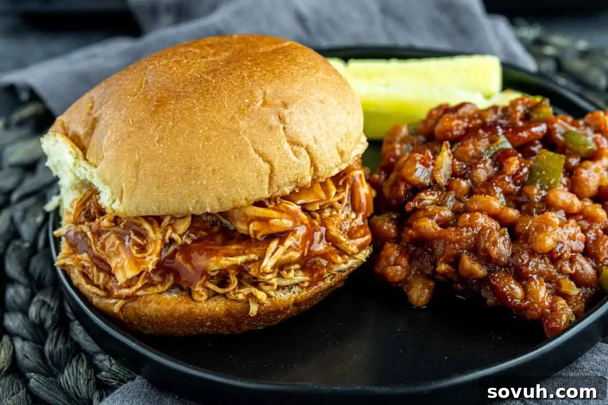 A delicious shredded barbecue chicken sandwich on a black plate, served with a side of baked beans and a crisp pickle slice. The chicken is glistening with sauce, promising a tender and flavorful bite.