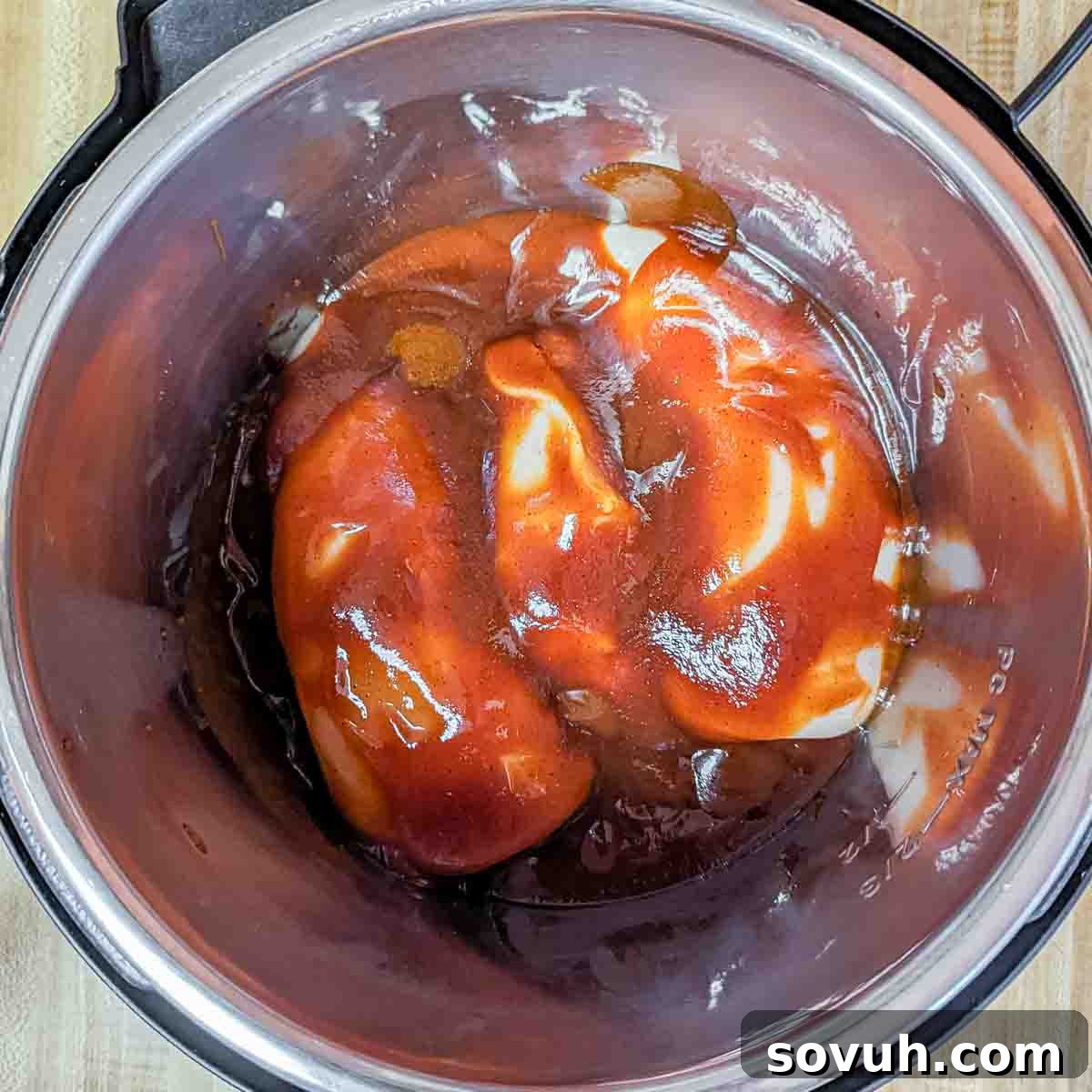 Two raw chicken breasts generously coated in barbecue sauce inside a stainless steel slow cooker, glistening and ready for cooking.