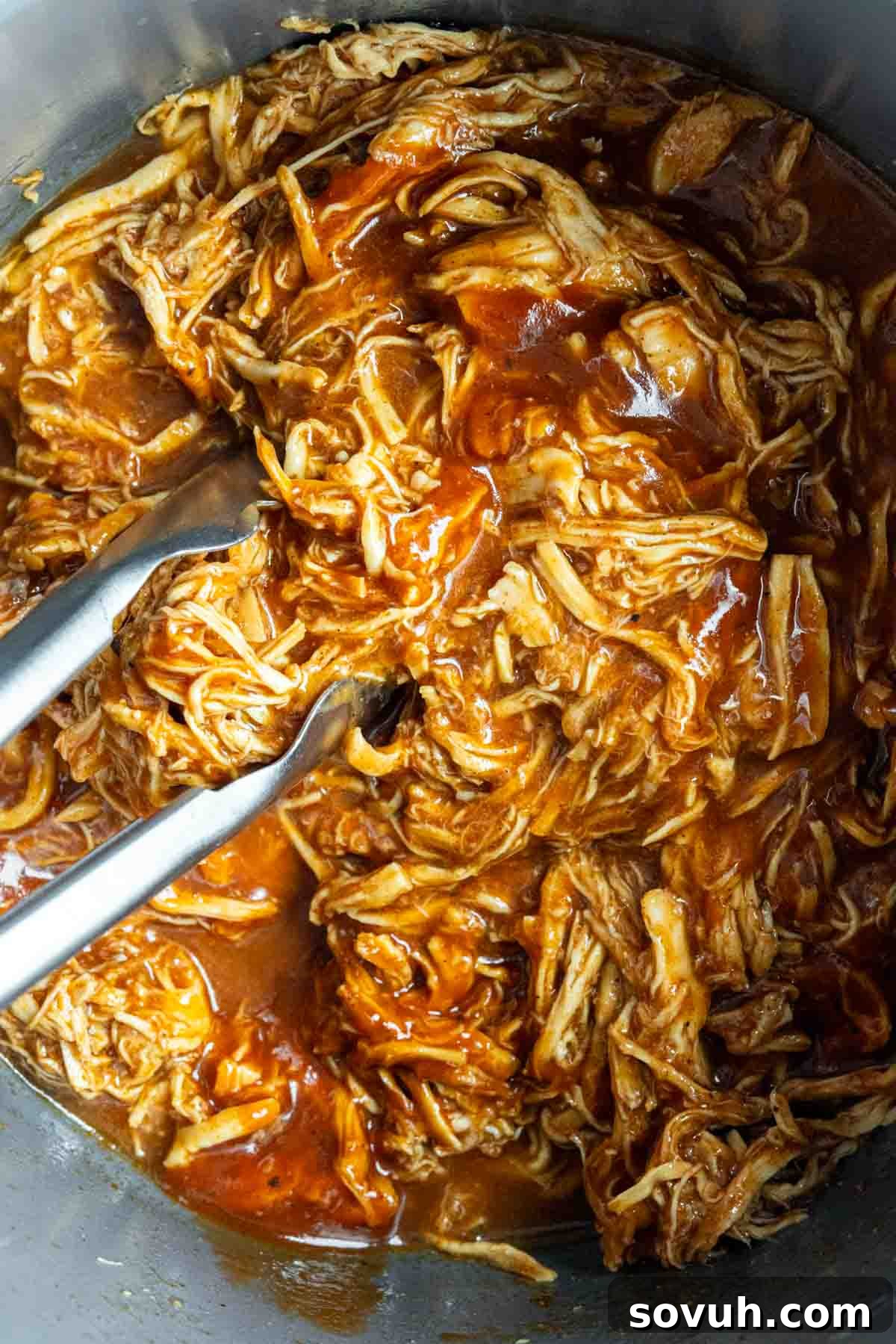 A close-up of tender, shredded barbecue chicken being mixed with tongs in a metal pot, highlighting its rich, reddish-brown sauce coating every strand of chicken.