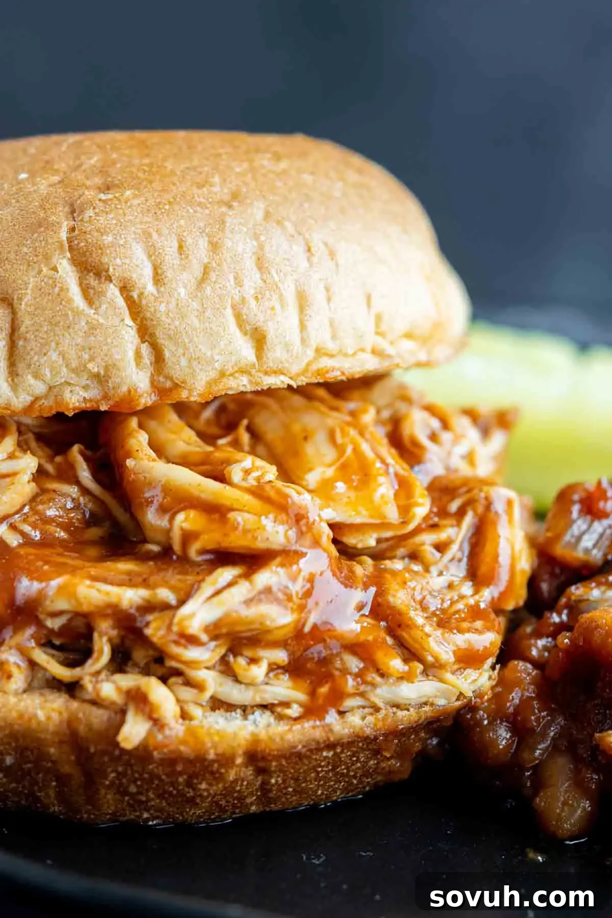 A mouth-watering barbecue chicken sandwich, featuring shredded chicken in a glossy sauce, served on a bun with a side of baked beans and a refreshing pineapple slice.