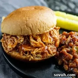A close-up of a barbecue pulled chicken sandwich on a black plate, accompanied by baked beans and pickle spears, highlighting the saucy, tender chicken.