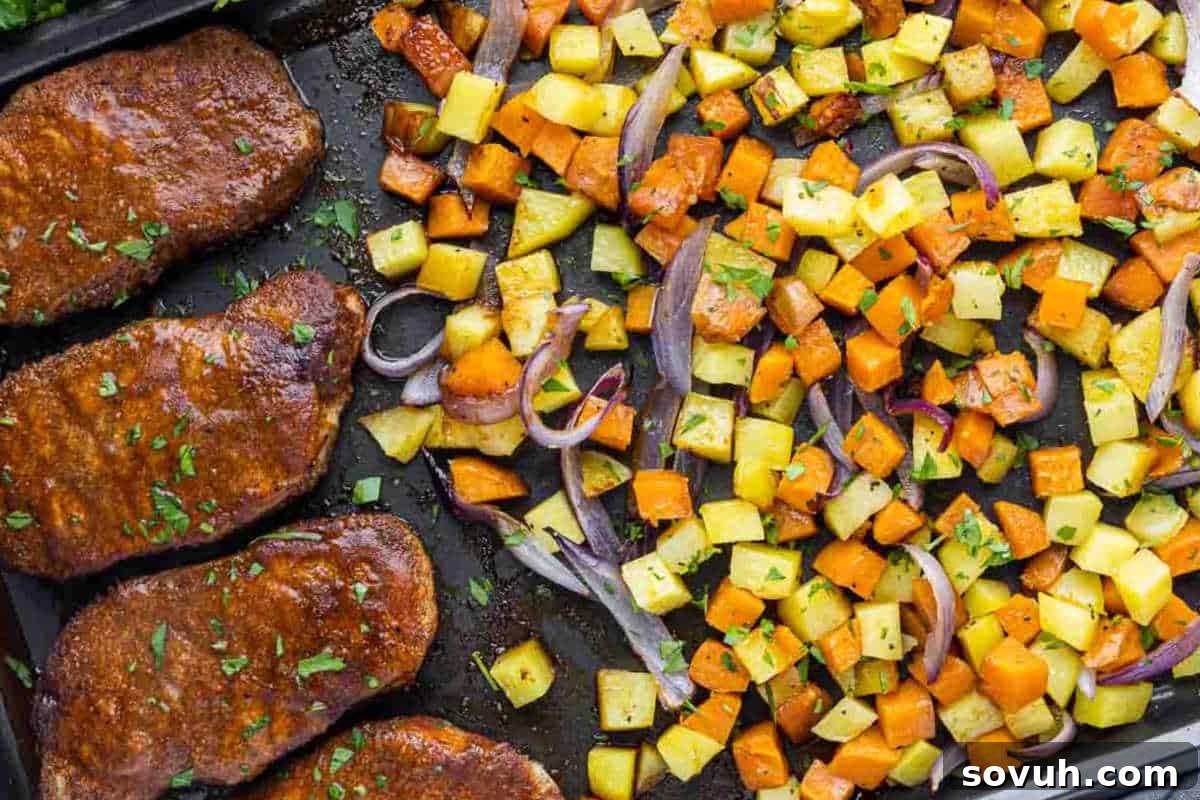 A baking sheet with four cooked pork chops on the left, seasoned with herbs, and diced roasted vegetables including sweet potatoes, yellow potatoes, and red onions on the right.