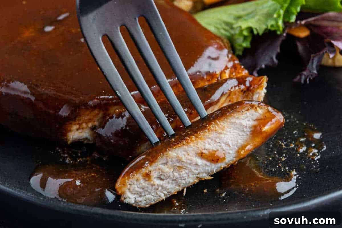 A fork holding a slice of barbecued meat with a glossy sauce on a black plate, accompanied by a side of greens.