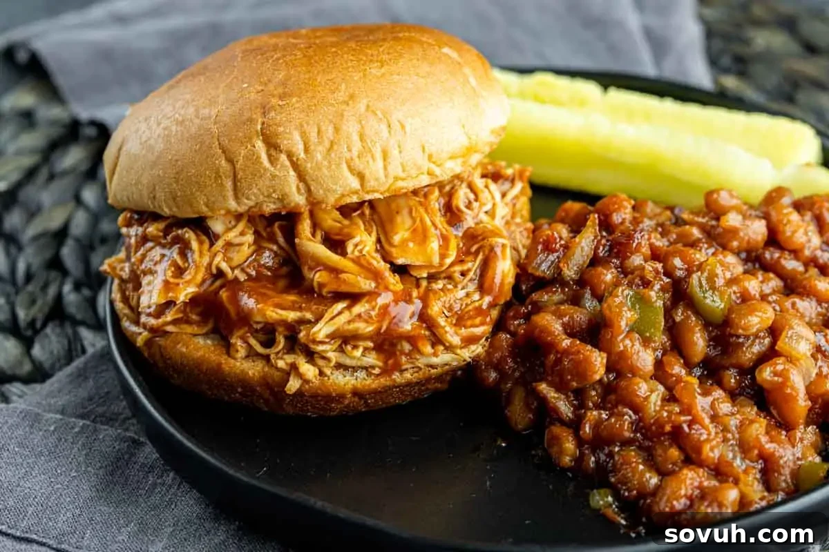 A pulled chicken sandwich with barbecue sauce is served on a black plate with a side of baked beans and pickle spears.