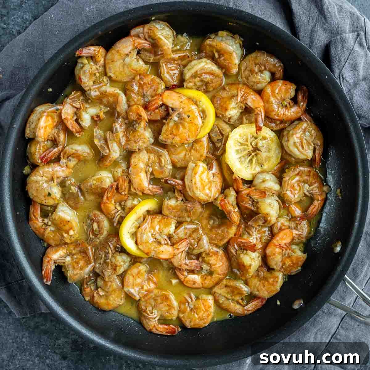 Overhead view of a skillet filled with cooked New Orleans BBQ Shrimp in a lemon butter sauce, garnished with lemon slices.