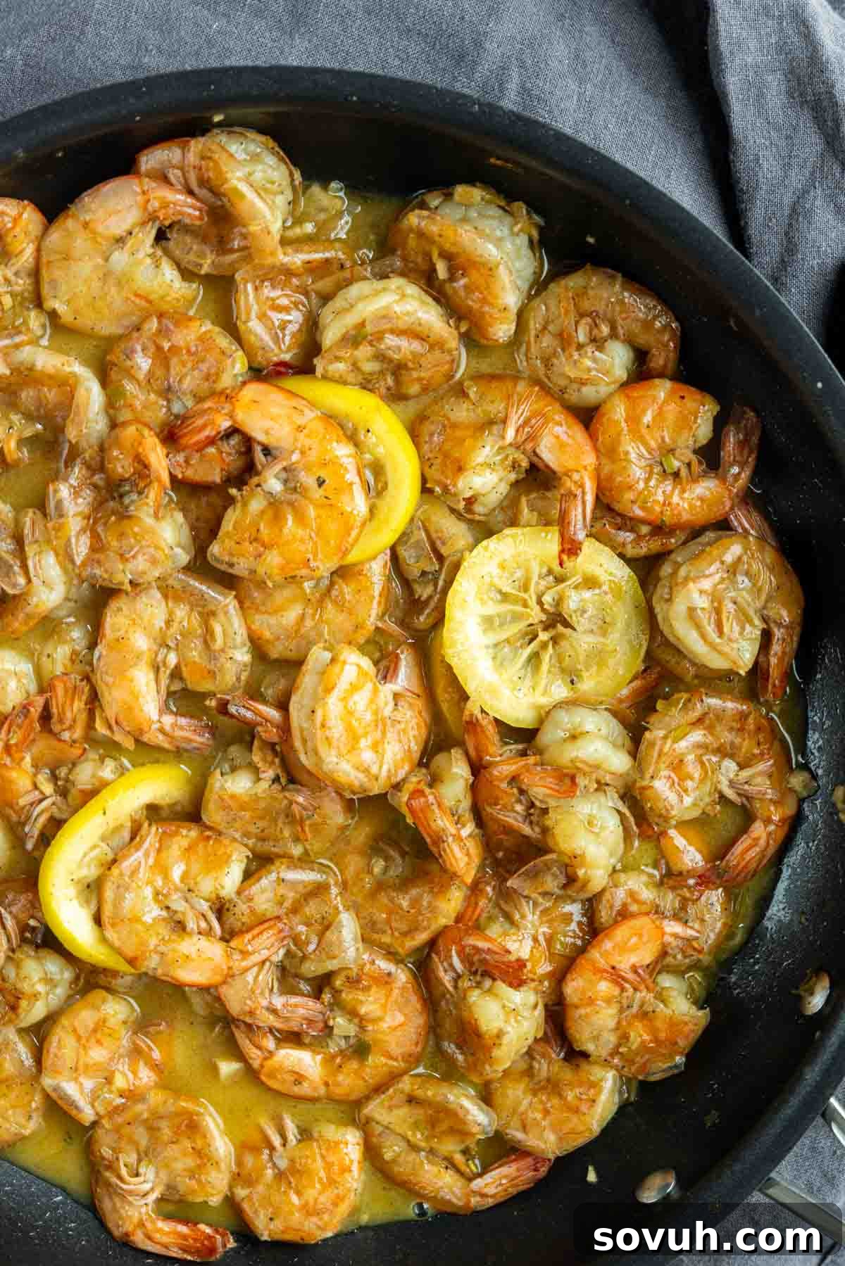 A skillet filled with cooked New Orleans BBQ Shrimp in a lemon garlic butter sauce, garnished with slices of lemon.