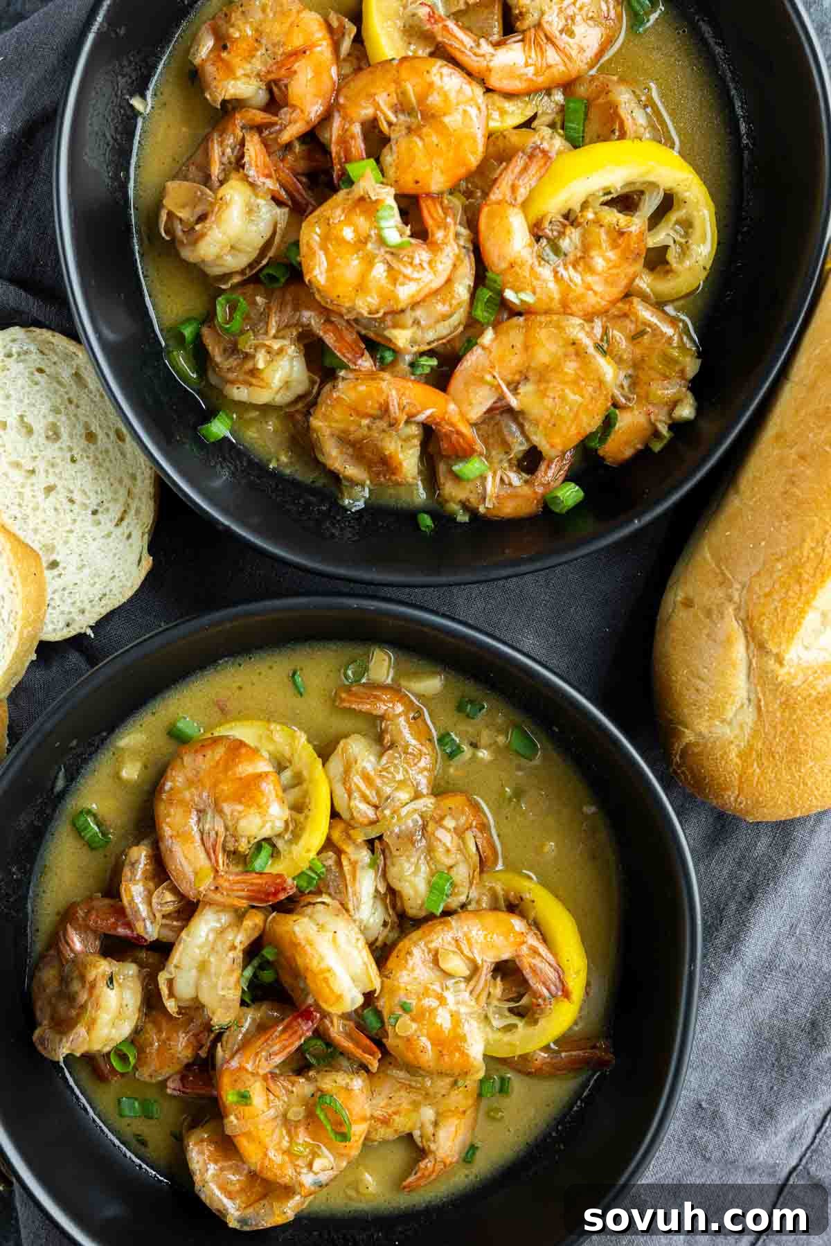 Two bowls of New Orleans BBQ Shrimp with lemon slices, garnished with green onions. Bread slices and a whole roll are next to the bowls.