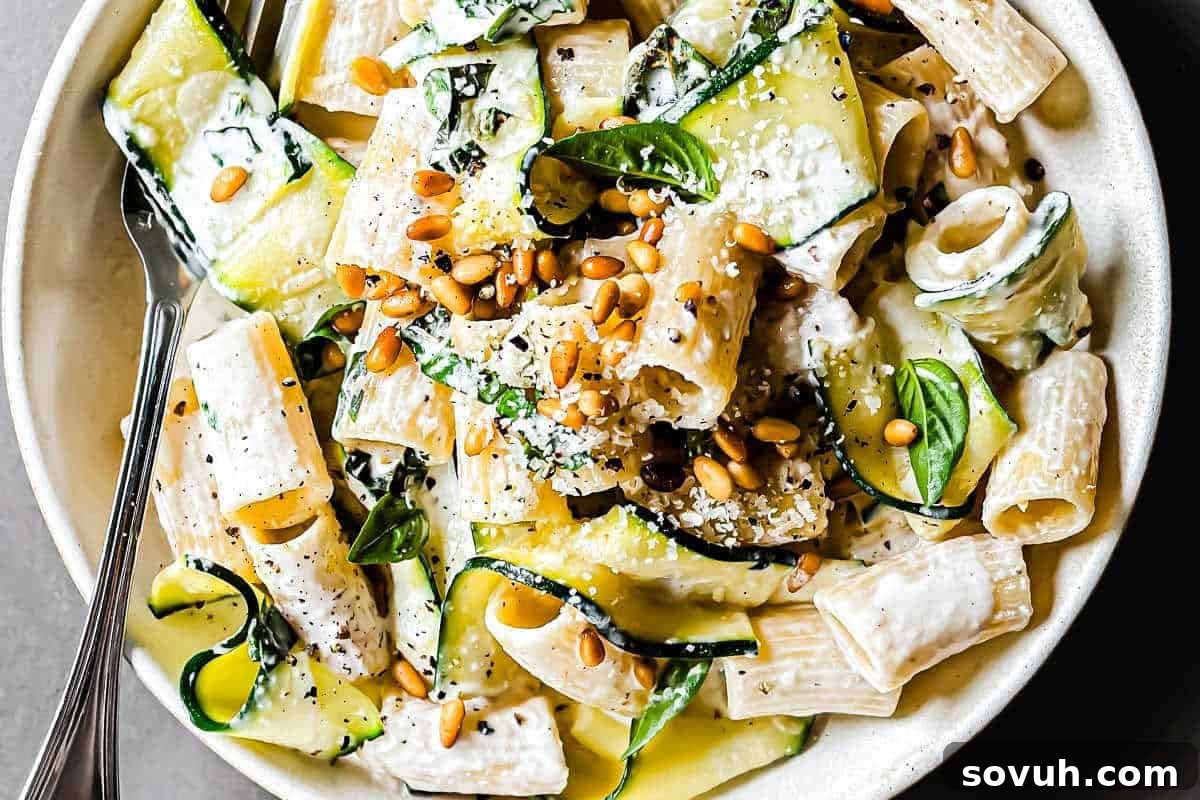 A bowl of pasta with creamy sauce, zucchini slices, fresh basil, and pine nuts, garnished with grated cheese and black pepper. A fork rests on the side of the bowl.