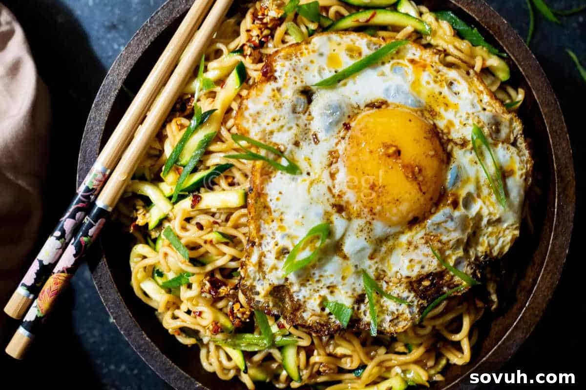 A bowl of stir-fried noodles topped with a fried egg, garnished with sliced green onions and served with chopsticks.