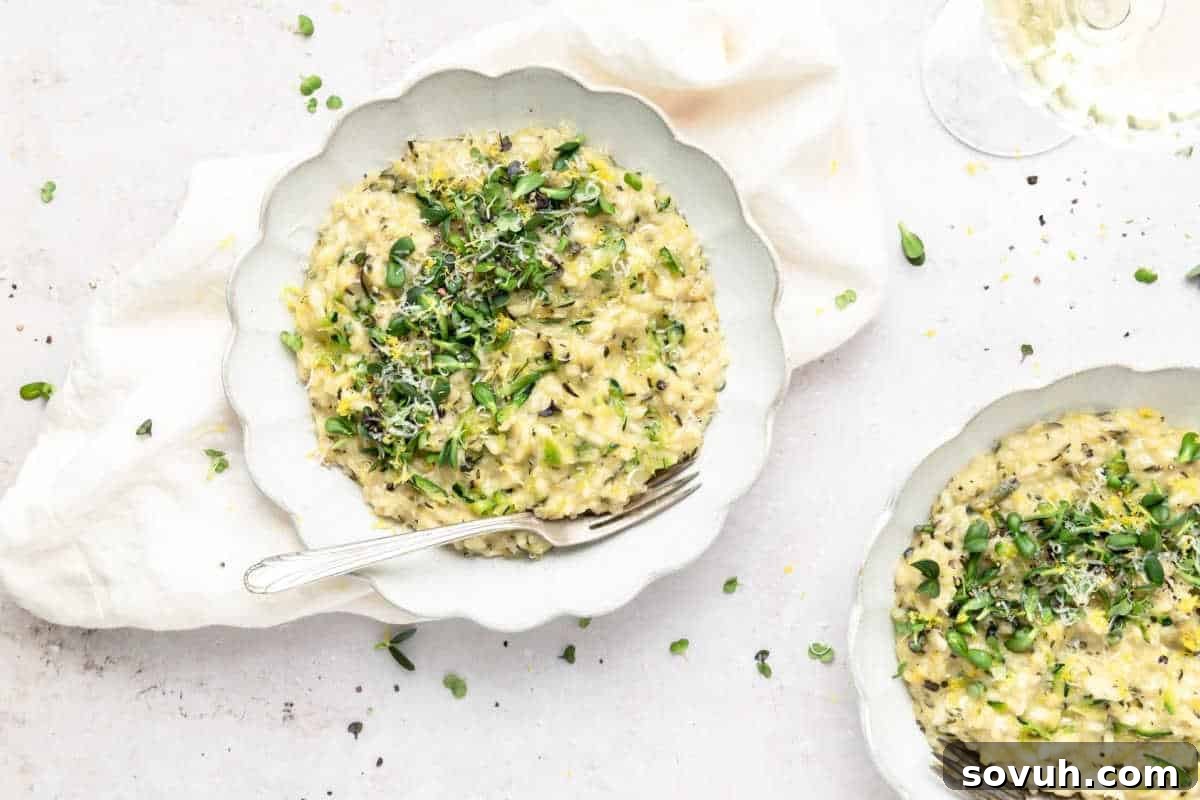 A bowl of zucchini risotto garnished with microgreens and grated cheese, placed on a white cloth. Another bowl of the same dish and a glass of white wine are partially visible.