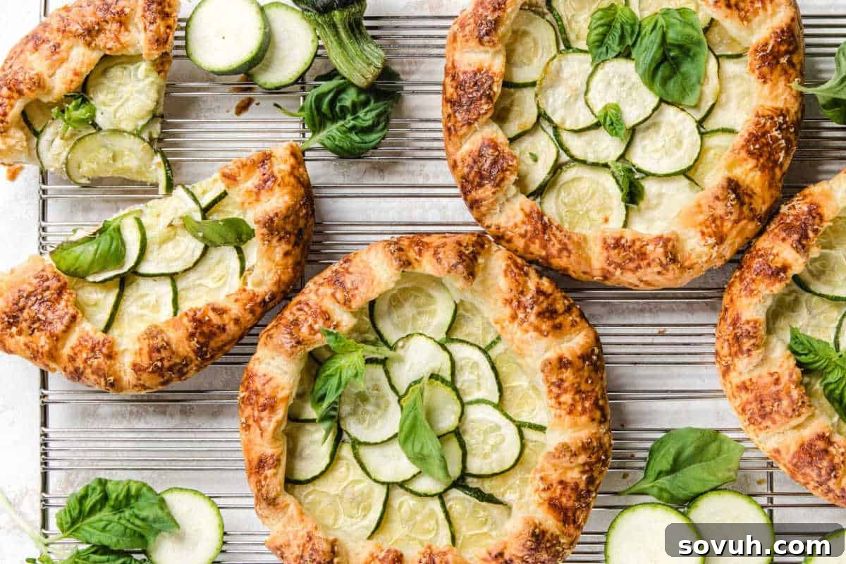 Four zucchini galettes on a cooling rack, each topped with thinly sliced zucchini and fresh basil leaves. Some additional basil leaves are scattered around the rack.