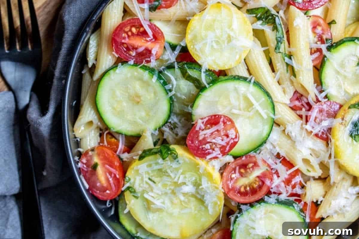 Close-up of a bowl of pasta salad with penne, zucchini slices, cherry tomatoes, and grated cheese on top. A fork and napkin are placed beside the bowl.
