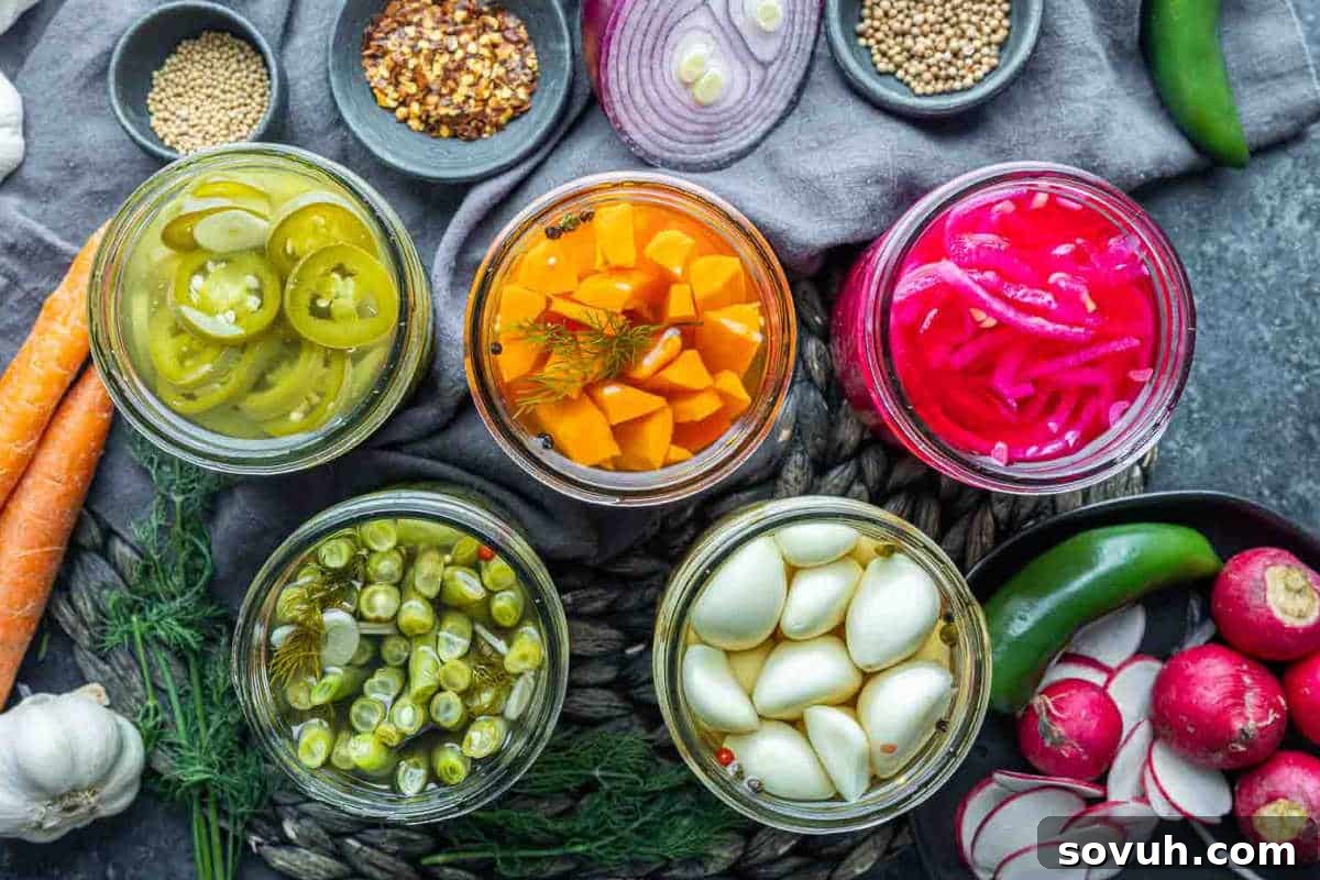 Five jars of pickled vegetables including jalapeños, carrots, red onions, green beans, and garlic, surrounded by spices, herbs, radishes, garlic, carrots, a jalapeño, and red onion on a gray surface.