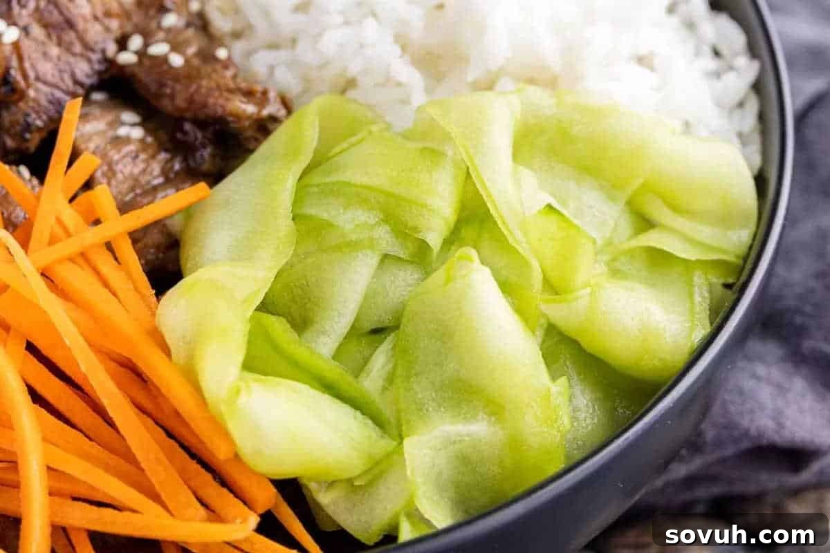 A bowl containing shredded orange carrots, sliced green vegetables, cooked meat pieces with sesame seeds, and white rice.