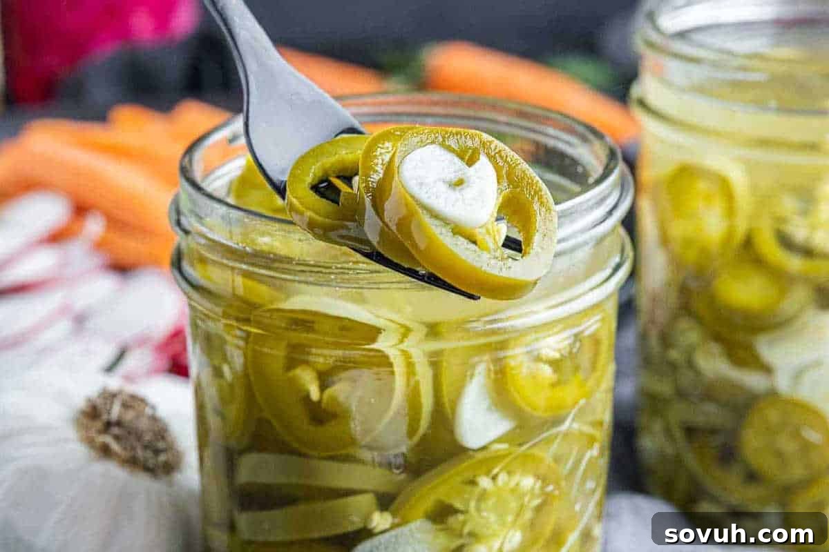 A fork holding pickled jalapeno slices above a jar filled with them. Other vegetables are blurred in the background.