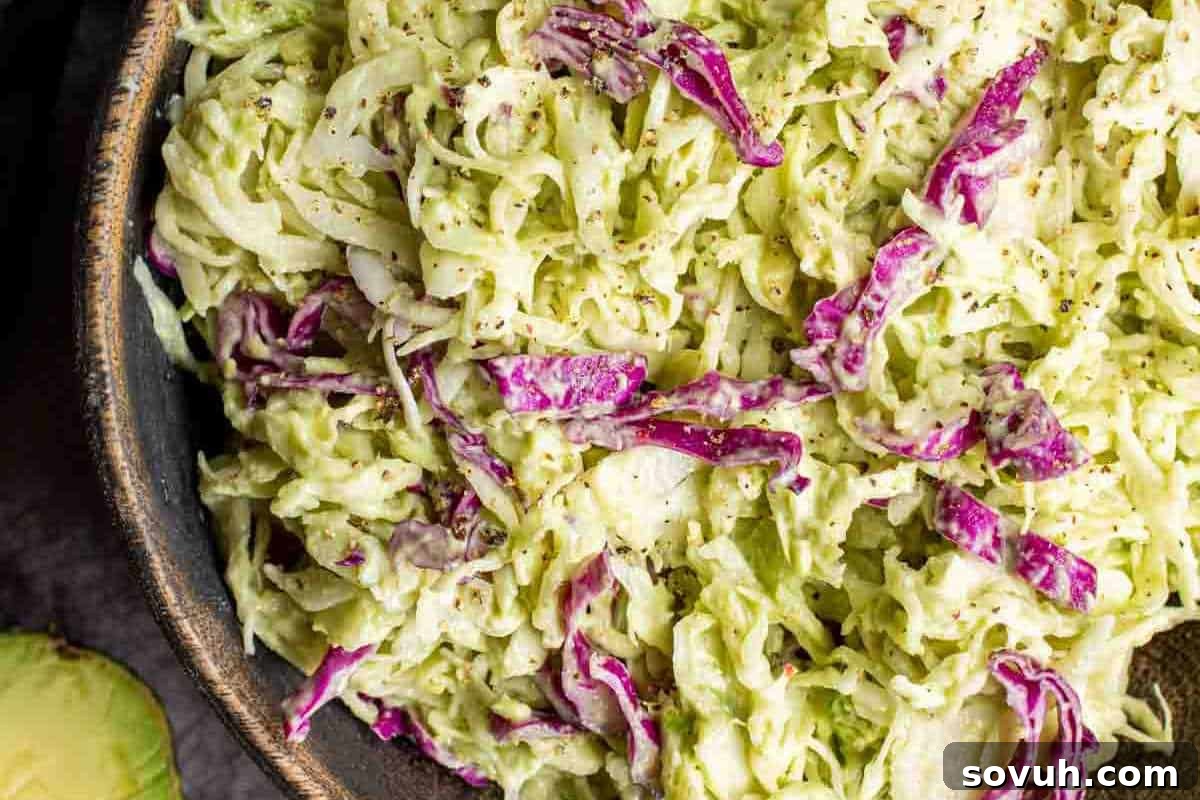 A close-up of a vibrant bowl filled with creamy coleslaw, featuring shredded green and purple cabbage mixed with a rich avocado dressing and sprinkled with black pepper.