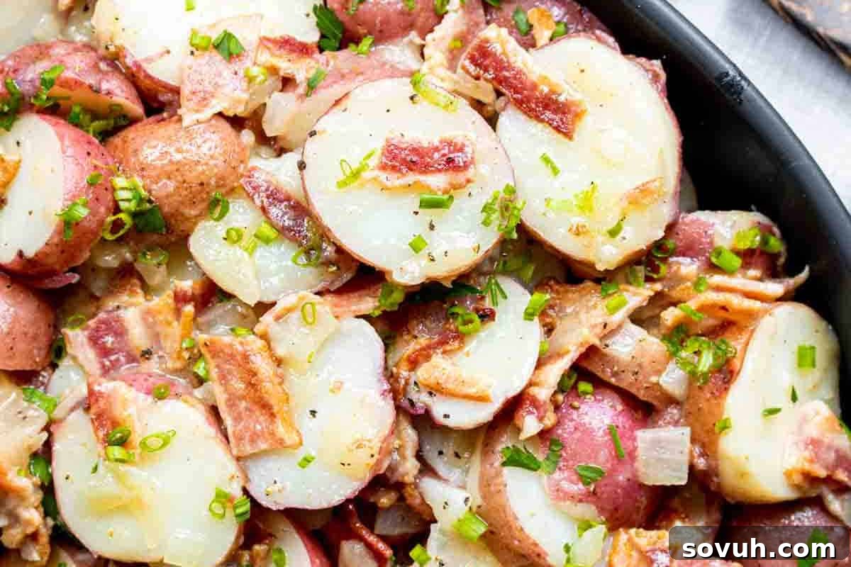 A comforting dish of perfectly sliced red potatoes generously topped with crispy bacon bits, sautéed onions, and a sprinkle of fresh green herbs.