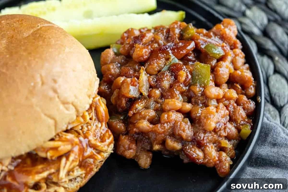 A close-up of a plate featuring a barbecue chicken sandwich on a bun, with a generous side of classic baked beans garnished with green peppers, and pickle spears in the background.