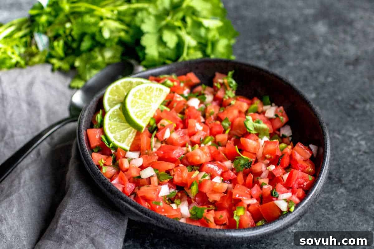 A bowl of pico de gallo garnished with lime slices, placed on a gray surface with fresh cilantro in the background and a spoon on a napkin beside it.