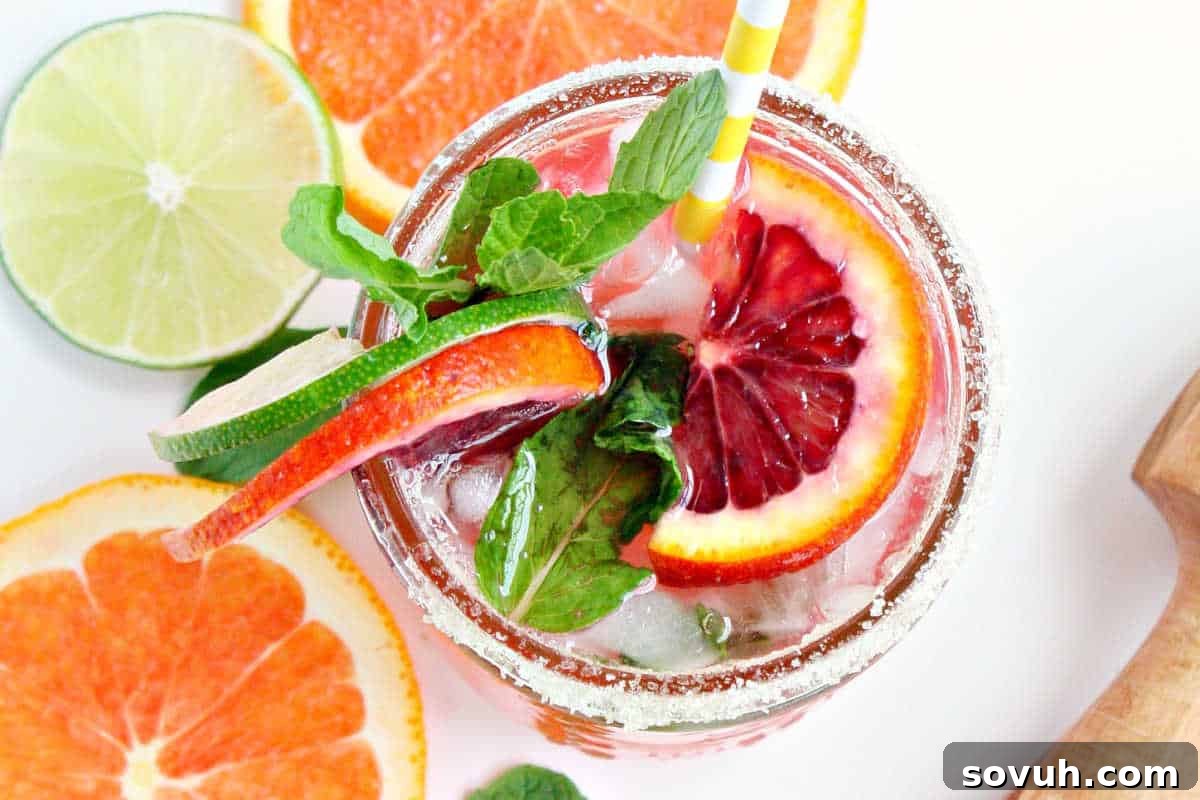 A glass of refreshing cocktail garnished with slices of blood orange, lime, mint leaves, and a striped straw, surrounded by cut citrus fruits.
