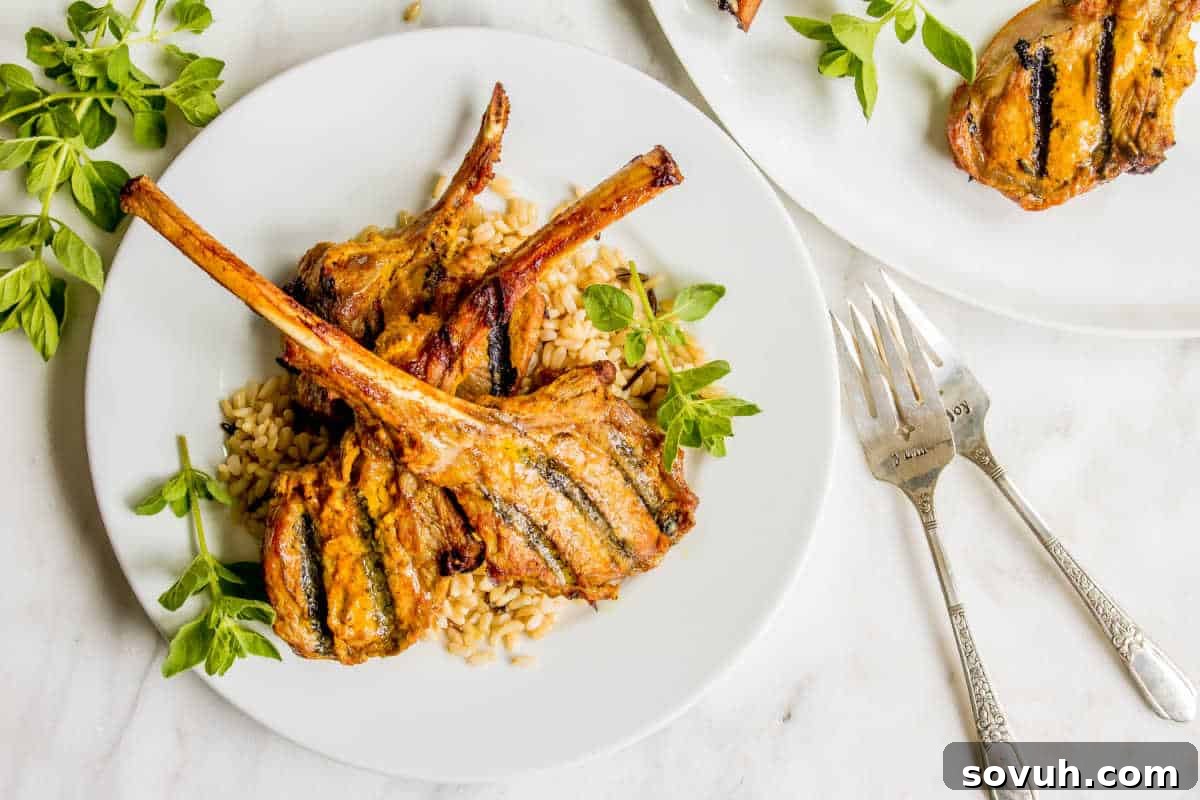 A plate with grilled lamb chops on a bed of orzo pasta, garnished with fresh herbs, next to a fork and knife on a marble surface.