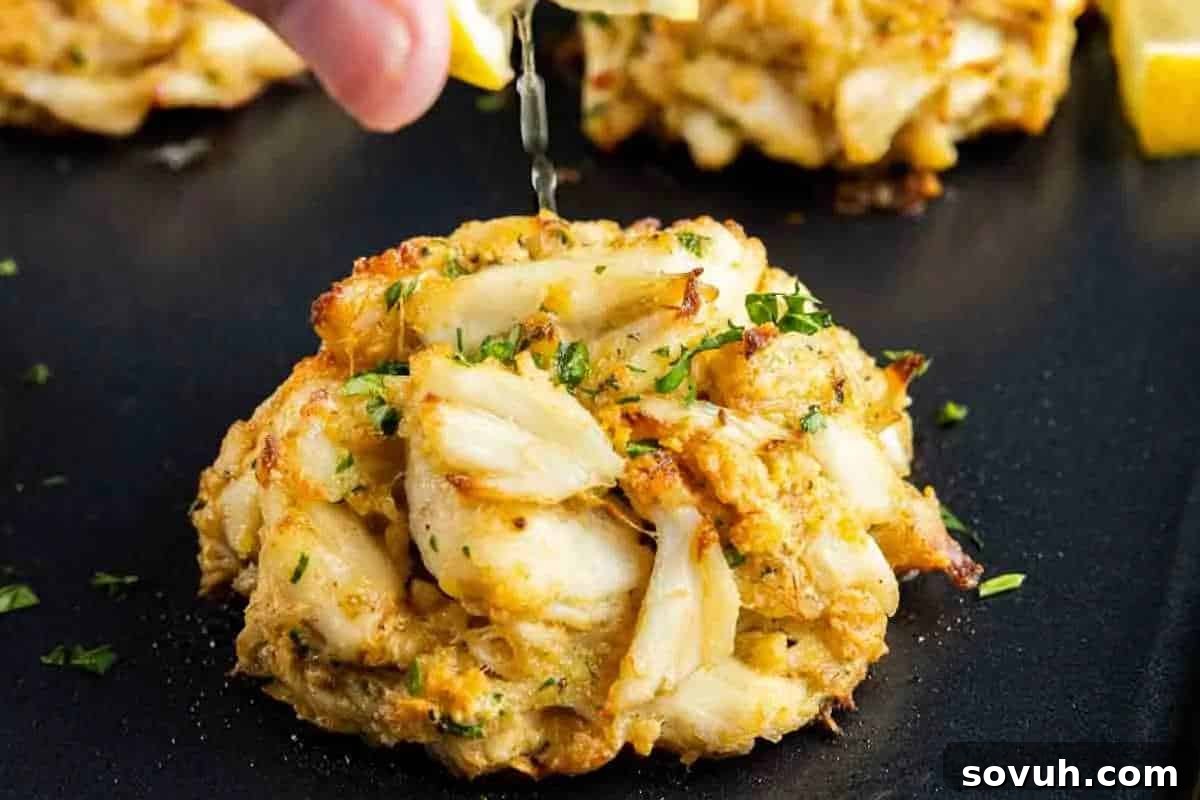 A hand squeezes a lemon wedge over a perfectly golden-brown Maryland crab cake, garnished with fresh herbs, resting on a sophisticated black surface. Two more delectable crab cakes blur in the background.