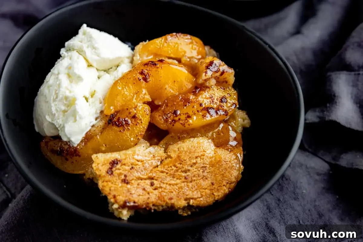 A black bowl with a serving of homemade peach cobbler, glistening with sweet juices and topped with a melting scoop of creamy vanilla ice cream, all presented on a rustic gray cloth background.