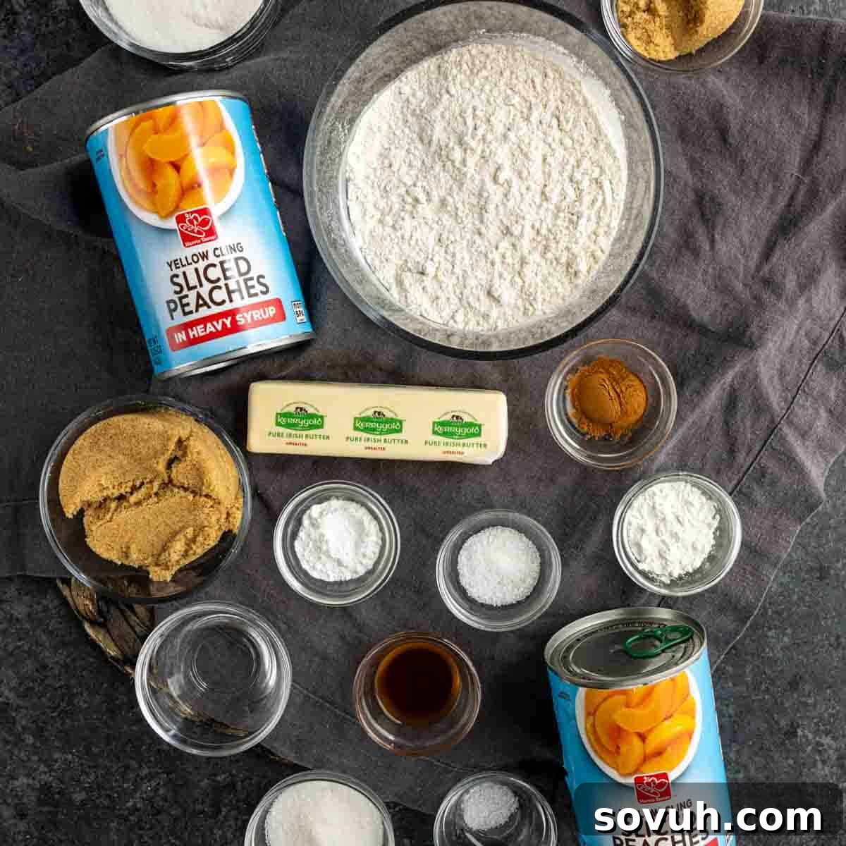 A vibrant display of all the essential ingredients needed for an easy peach cobbler, including glistening canned peaches, flour, brown and granulated sugars, butter, milk, vanilla extract, cornstarch, and aromatic cinnamon, all neatly arranged on a rustic wooden table.