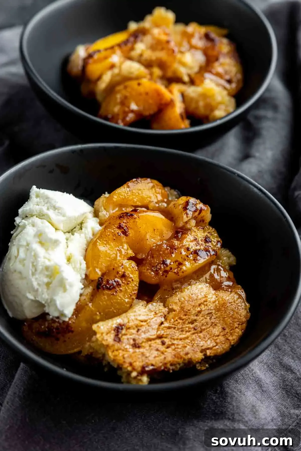 Two enticing bowls of golden peach cobbler served on a dark cloth; one bowl includes a generous scoop of melting vanilla ice cream, and both showcase the sweet caramelized peaches and crisp, golden crust.