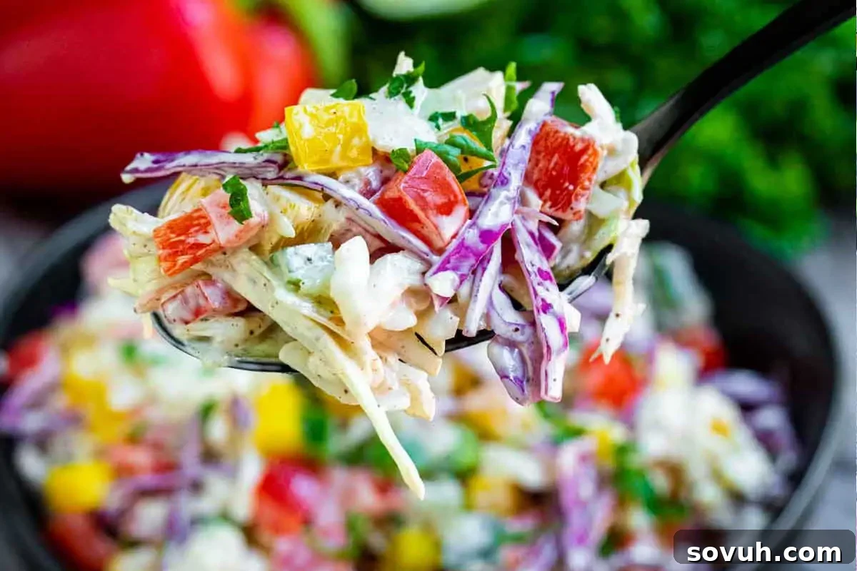 Close-up of a fork holding a colorful coleslaw with red, yellow, and green bell peppers, purple cabbage, and creamy dressing, with more coleslaw blurred in the background.