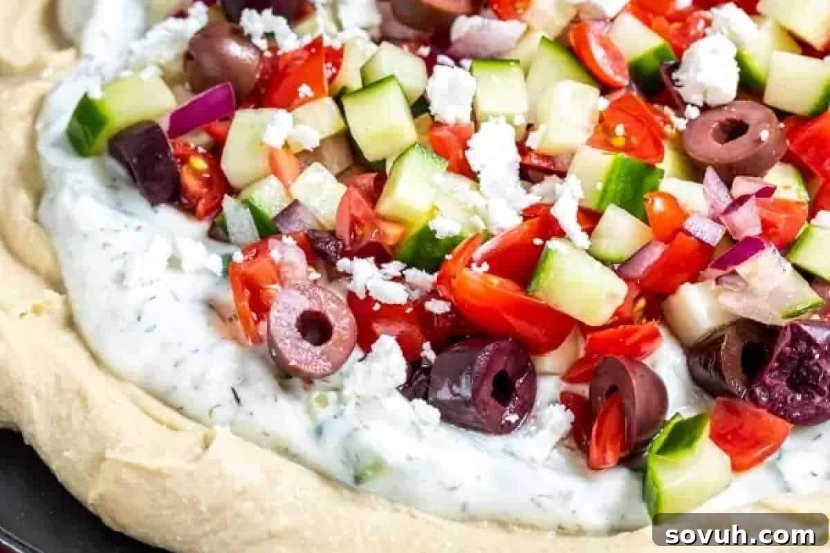 A close-up of a layered Mediterranean dip featuring chopped cucumbers, tomatoes, red onions, Kalamata olives, and crumbled feta cheese on a creamy base.