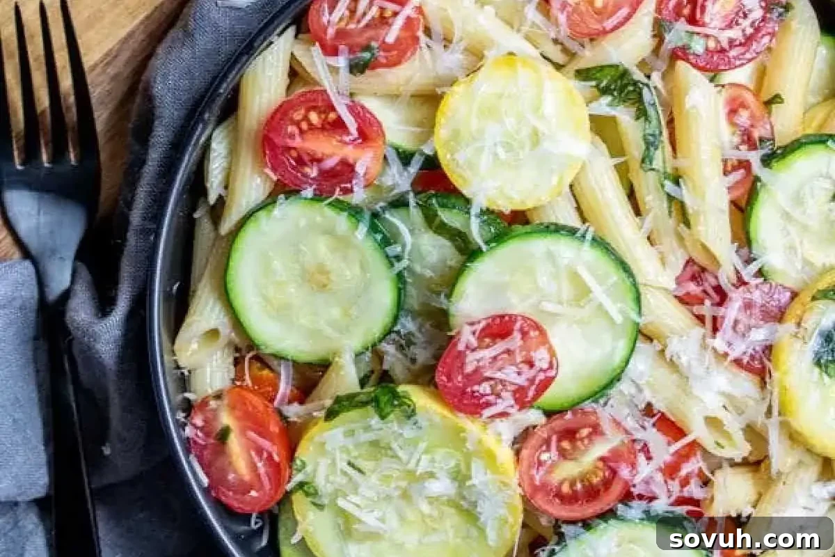 A close-up of a pasta salad featuring penne, cherry tomatoes, and slices of zucchini, topped with grated cheese and garnished with basil leaves.