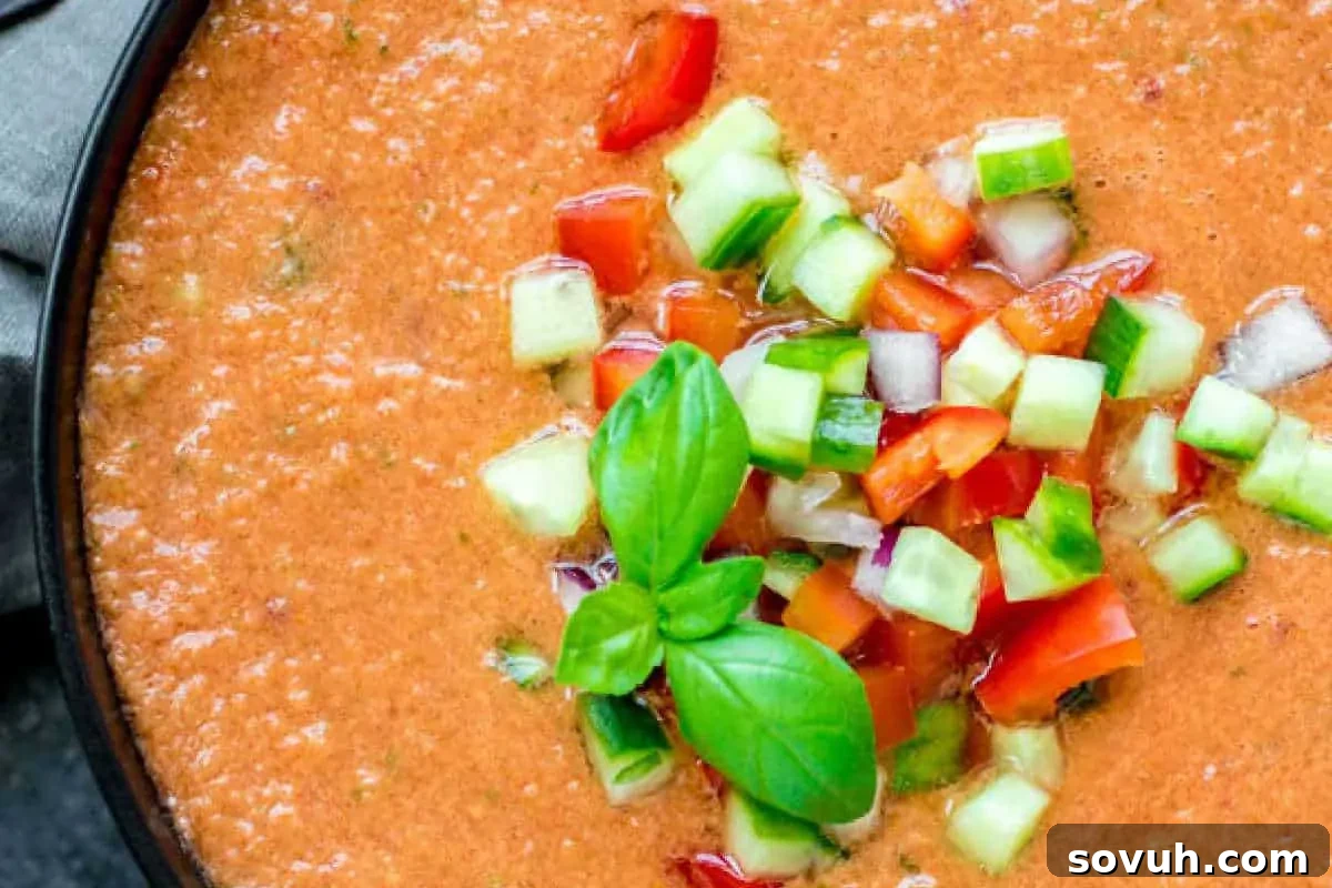 A bowl of gazpacho garnished with diced cucumber, red bell pepper, red onion, and fresh basil leaves.