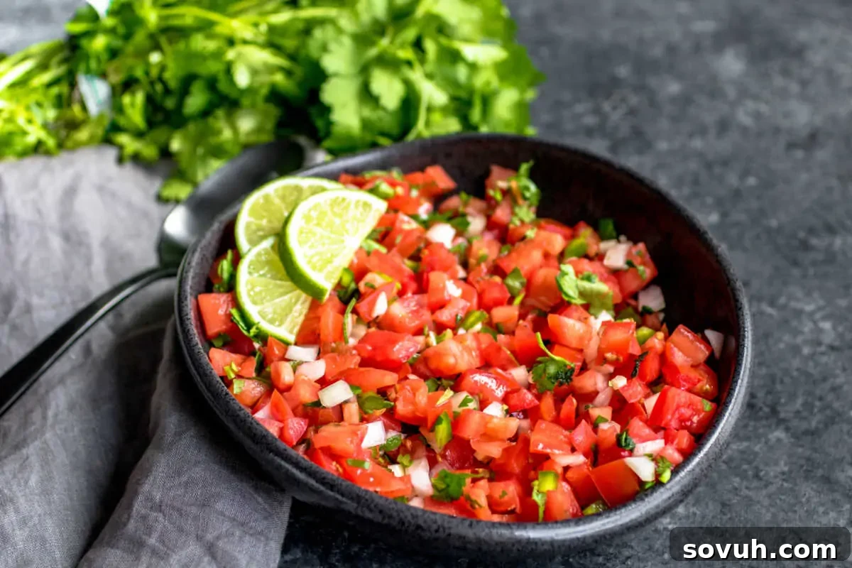 A bowl of fresh pico de gallo garnished with sliced lime, placed on a gray surface with a bunch of cilantro and a spoon in the background.