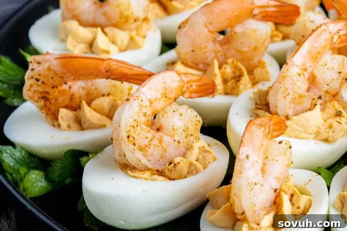 A beautifully arranged platter of deviled eggs, each half topped with a perfectly seasoned cooked shrimp and garnished with fresh green herbs. The background features leafy greens, suggesting a sophisticated presentation.