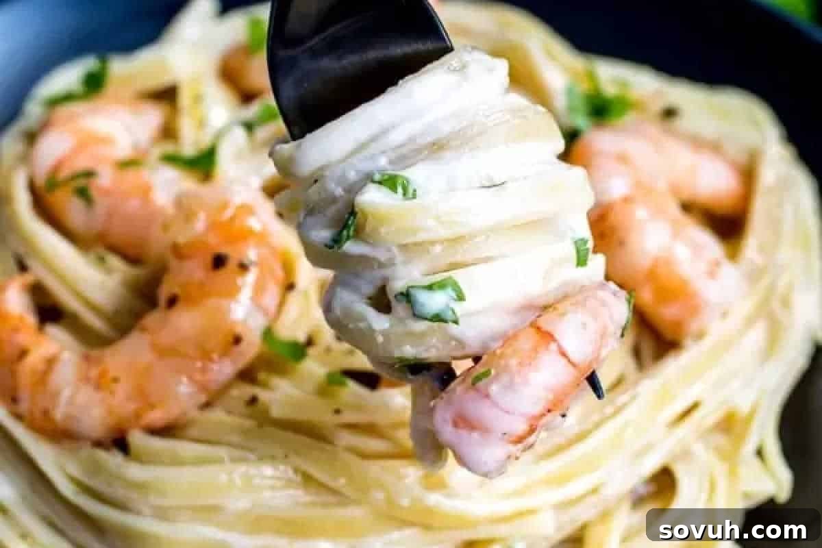 A fork expertly twirls creamy fettuccini pasta with succulent shrimp, garnished generously with fresh chopped herbs. In the background, a serving bowl overflowing with more Shrimp Alfredo Pasta suggests a hearty and inviting meal.