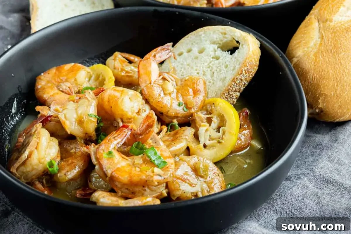 A deep black bowl brimming with authentic New Orleans BBQ Shrimp, showcasing plump, perfectly cooked shrimp bathed in a rich, tangy butter sauce. Bright lemon slices and vibrant green onions garnish the dish, with a slice of crusty French bread on the side, ready for dipping.