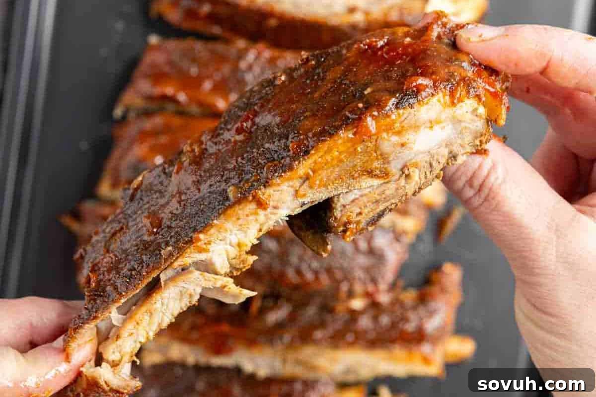 Hands holding a piece of barbecued ribs with a dark, glossy sauce, in front of a platter of more ribs.